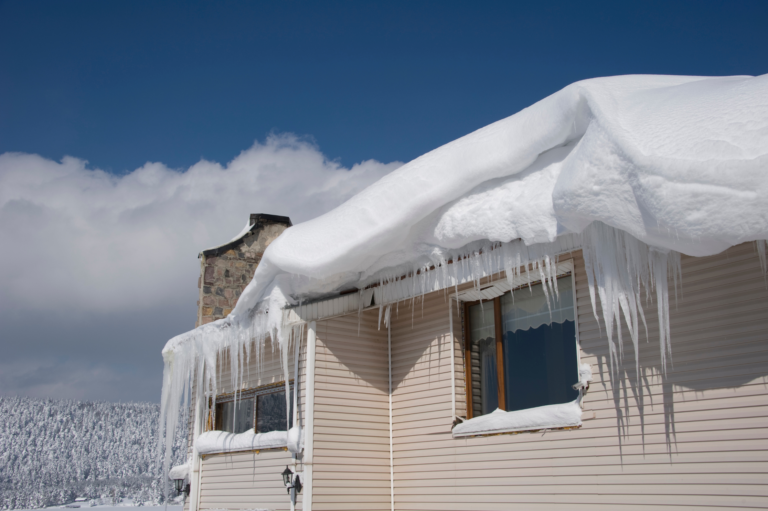 A residential home after a heavy snowfall, showcasing the real-life effects of ice damming on the roof. The thick blanket of snow on the roof overhangs the edge, bending downwards due to its weight. Large icicles have formed along the eaves, indicating that melting snow has refrozen, a sign of potential ice dams. The backdrop features a bright blue sky and a forest densely coated in snow, highlighting the wintry conditions.