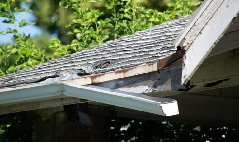 Close-up of a damaged roof with missing shingles, broken gutters, and wood rot, illustrating storm and wind damage.