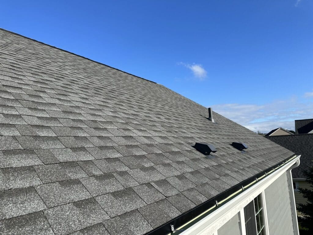 A close-up view of a residential roof with gray asphalt shingles under a clear blue sky. The roof features two black attic vents and a metal exhaust pipe, with white gutters along the roofline. Neighboring houses with similar roofing are visible in the background.